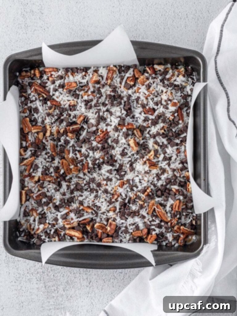 Golden Coconut Dream Bars 5 A square pan filled with layered magic bar mixture, ready for baking. The layers of graham cracker crust, condensed milk, and toppings are visible.
