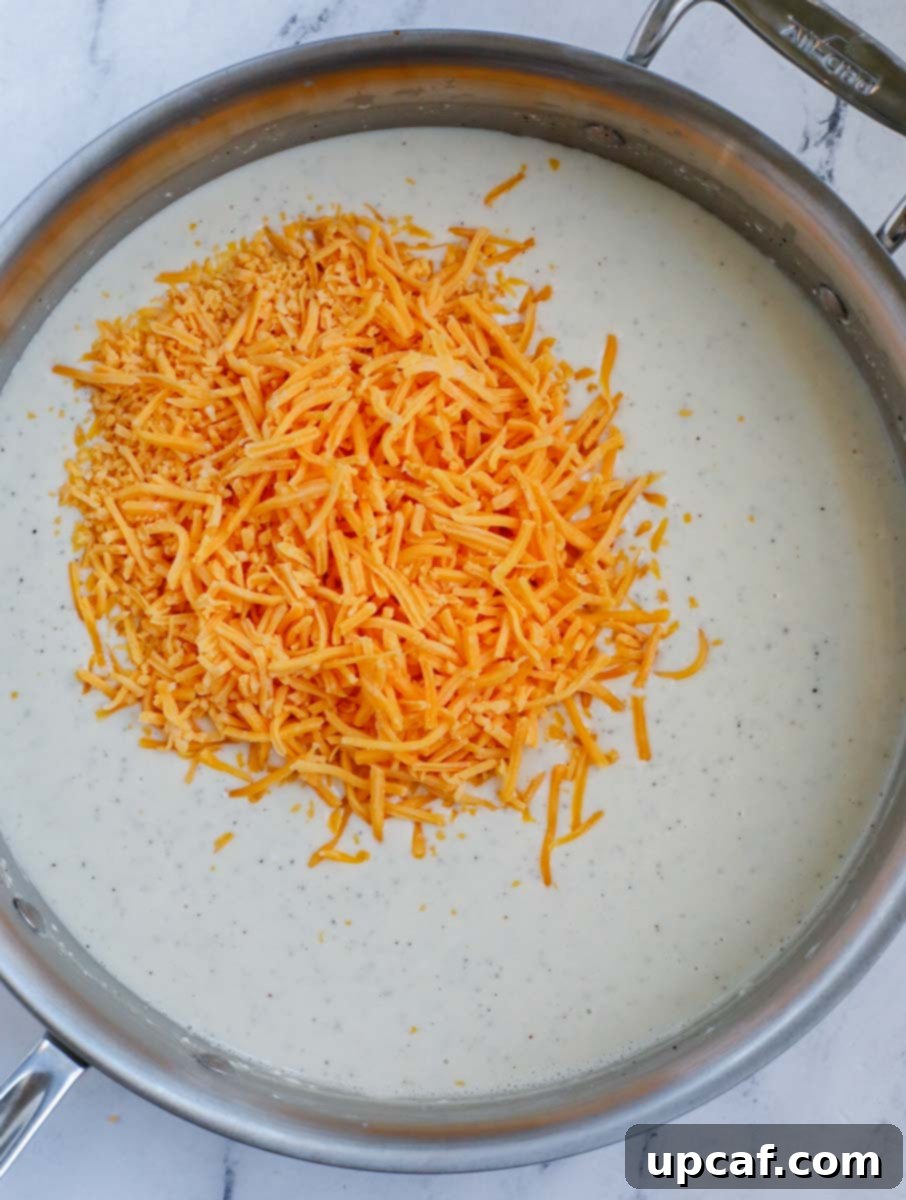 A silver skillet with creamy sauce and a heaping pile of shredded cheddar cheese waiting to be mixed in.