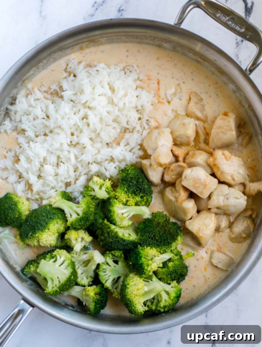 A silver skillet with a smooth creamy casserole sauce at the bottom, topped with cooked rice, chicken, and broccoli, ready for mixing.
