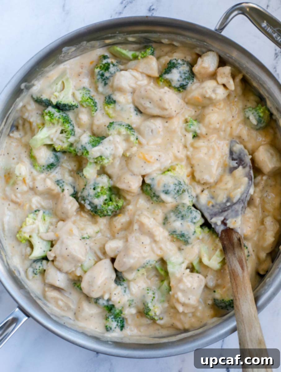 A silver skillet filled with a mixed chicken, broccoli, and rice casserole in a sauce, with a wooden spoon resting on a white counter.