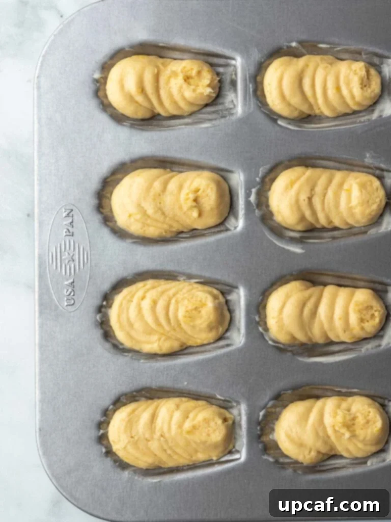 A close-up view of the delicate lemon Madeleine batter expertly piped into the individual shell-shaped cavities of a baking pan, poised for baking.