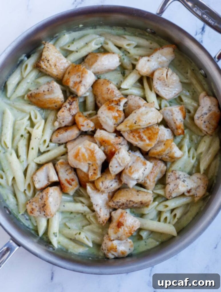 Cooked chicken cubes and penne pasta thoroughly mixed with a vibrant pesto sauce in a skillet.