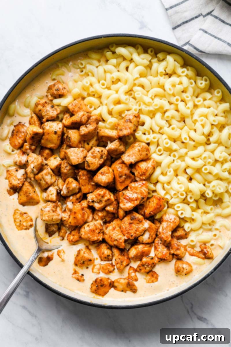 Cooked Cajun chicken and macaroni being added to the creamy cheese sauce in a skillet.