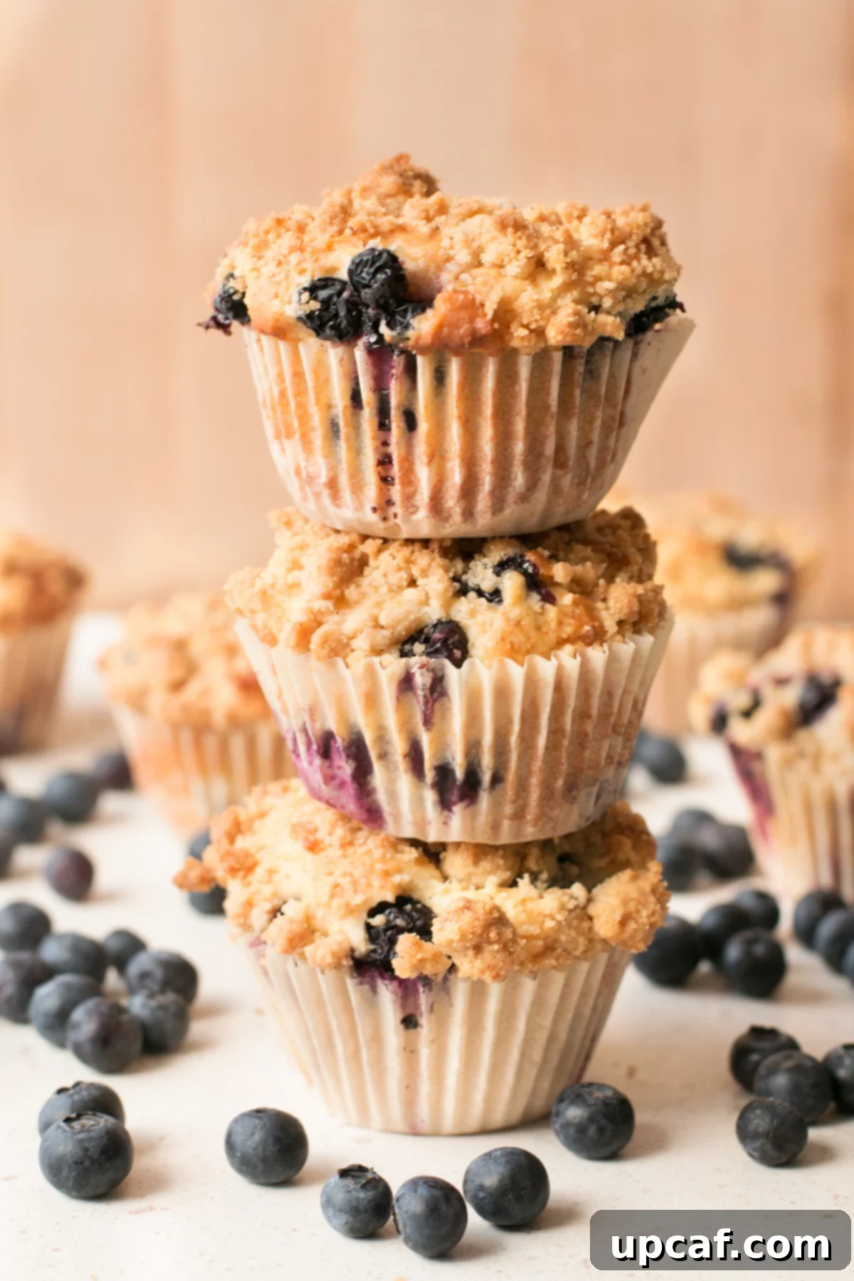 A stack of blueberry crumb muffins sitting on a white surface and surrounded by loose blueberries