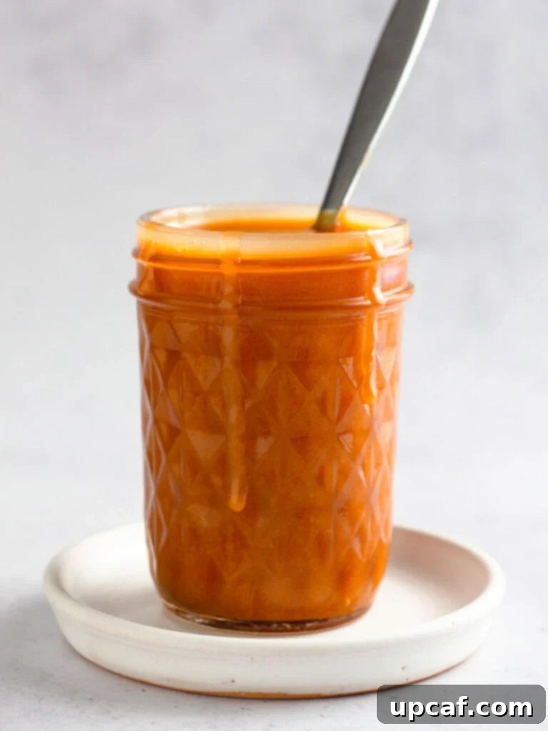 The freshly made caramel sauce poured into a clear mason jar, with a spoon resting in it, showcasing its smooth texture.