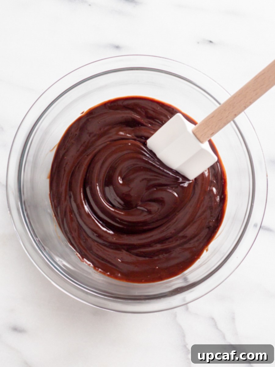A rubber spatula gently mixing melted chocolate and hot cream in a bowl, creating a smooth ganache.