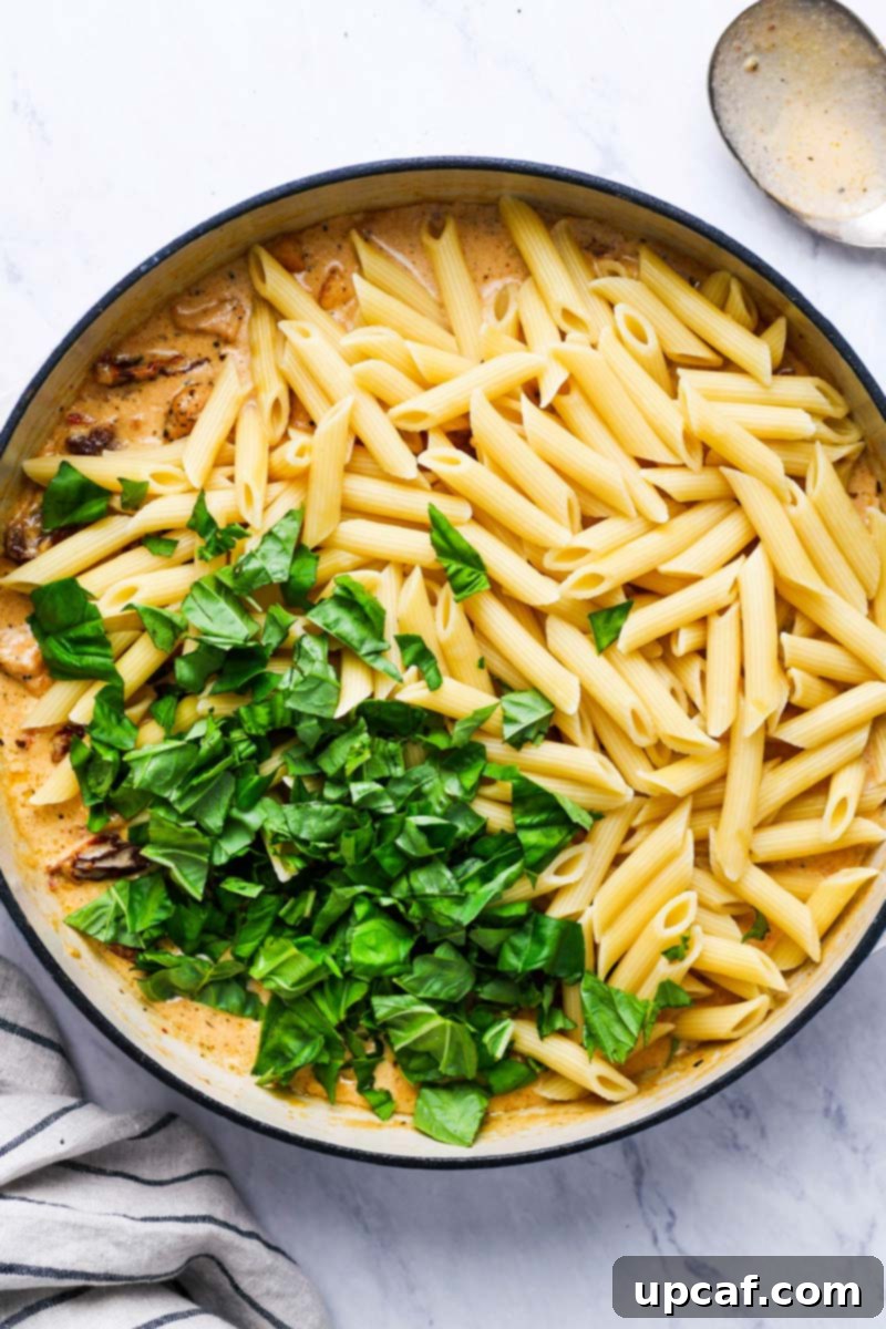 Adding pasta and fresh basil to the cream sauce to be coated, showcasing the vibrant colors.