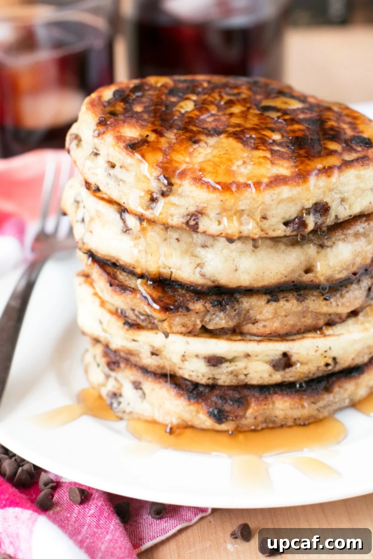 A stack of chocolate chip pancakes on a white plate drizzled with pancake syrup