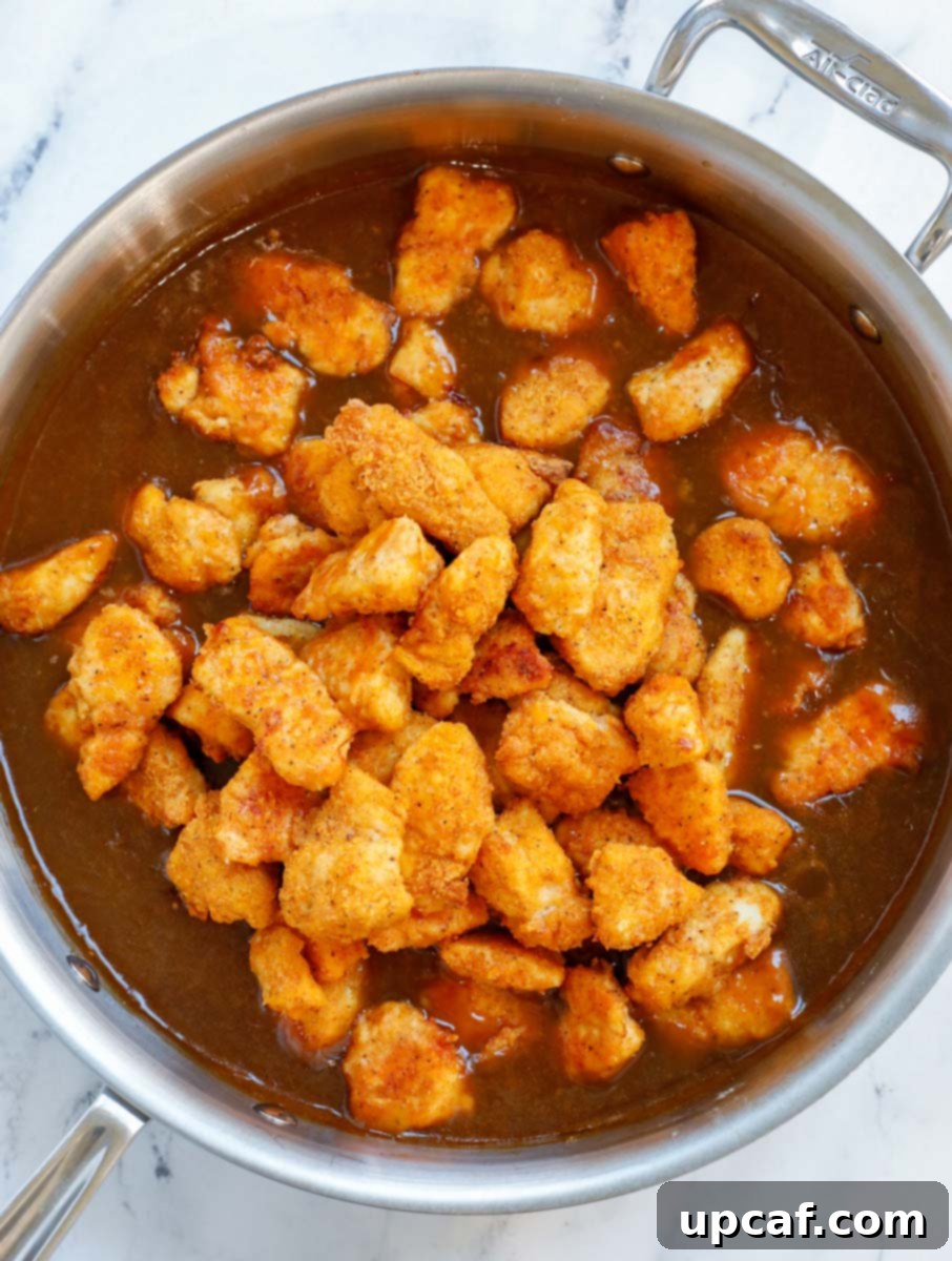Wholesome Orange Chicken 8 Adding freshly baked, crispy orange chicken pieces directly into the simmering sauce in a large pan.