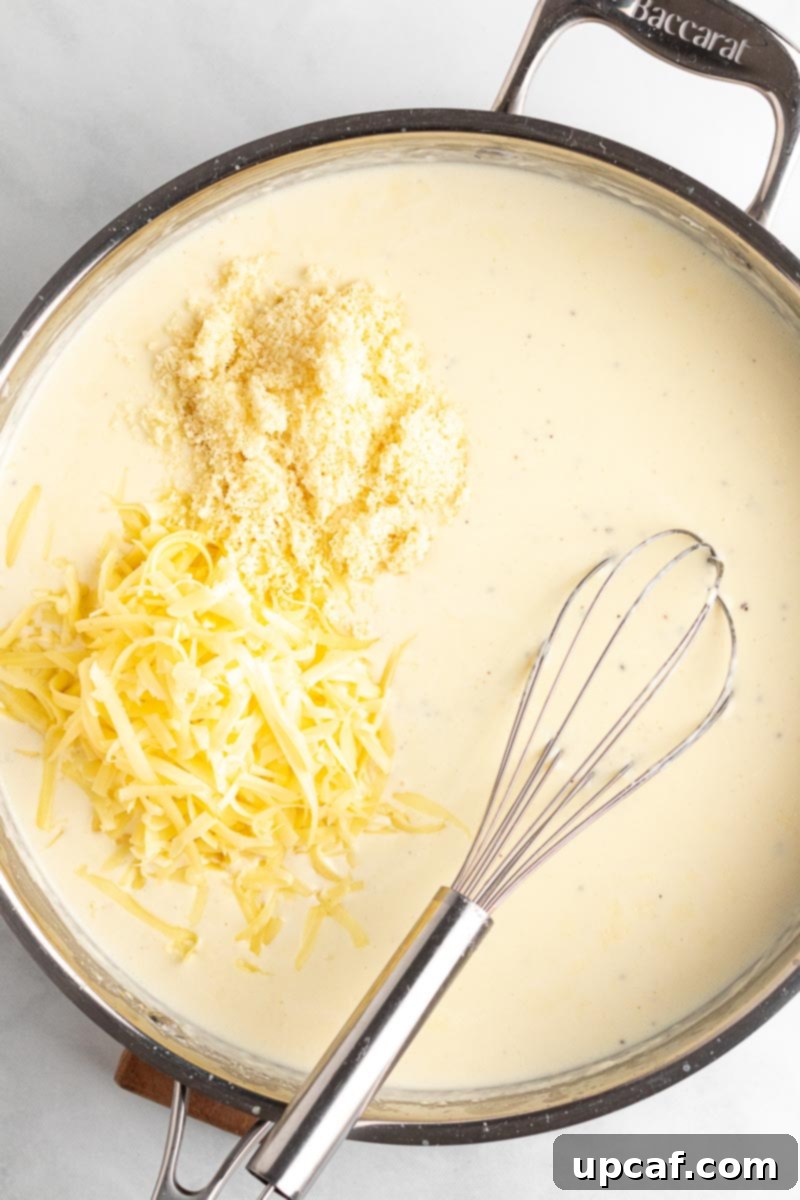 Freshly shredded sharp white cheddar and Parmesan cheeses being added to the simmering milk mixture in a skillet.