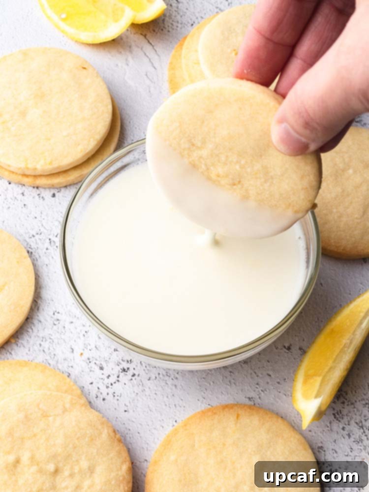 Tangy Lemon Glazed Shortbread 10 A shortbread cookie being dipped halfway into a bowl of lemon icing, showcasing the technique for achieving a perfectly coated half-moon design.