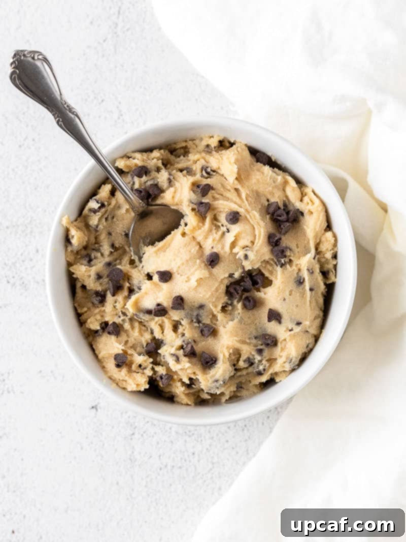 Irresistible Eggless Cookie Dough 6 A white bowl brimming with freshly made edible cookie dough, speckled with chocolate chips, and a spoon ready for tasting.