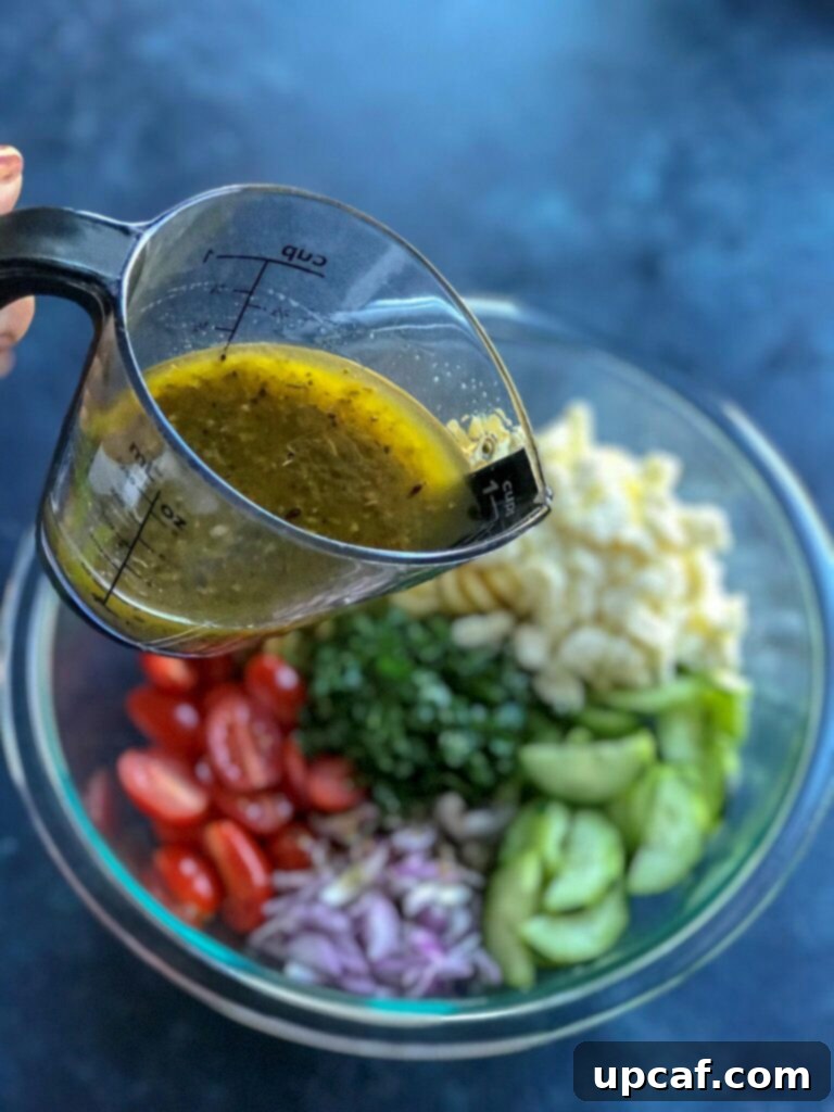 Vibrant Greek Pasta Salad 4 Close-up shot of lemon vinaigrette being poured over the fresh ingredients of Greek Pasta Salad, showing the process of dressing the salad.