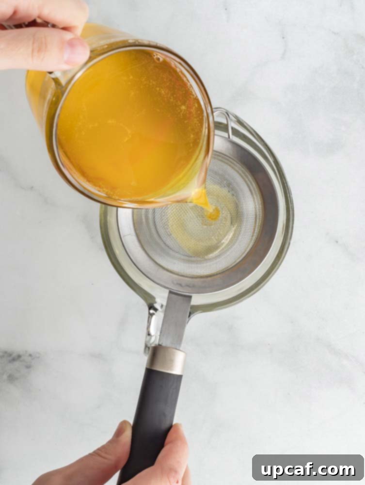 Freshly cooked ghee being strained through a fine-mesh sieve lined with cheesecloth into a glass jar.