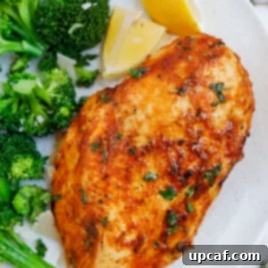 Close up of an air fryer rosemary chicken breast beside some broccoli and lemon wedges.