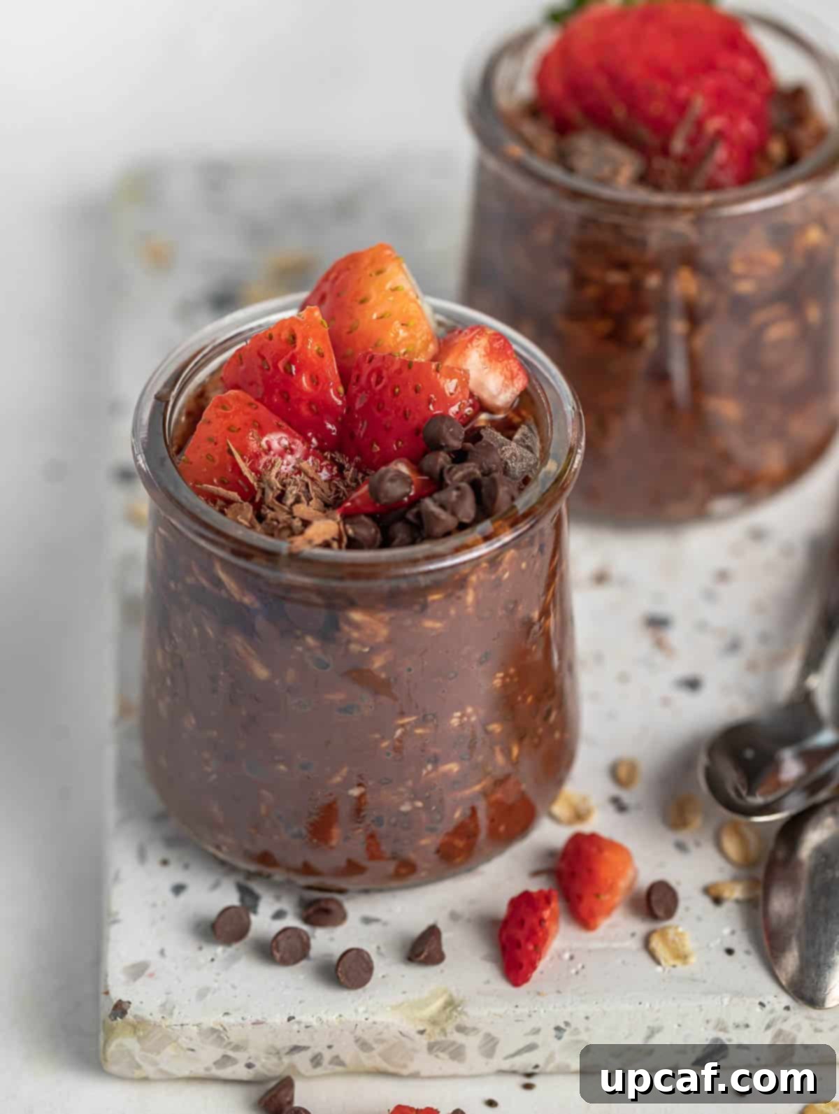 A vibrant display of two cups of chocolate overnight oats, garnished with fresh strawberries and chocolate chips, against a bright background, emphasizing their appetizing appeal.