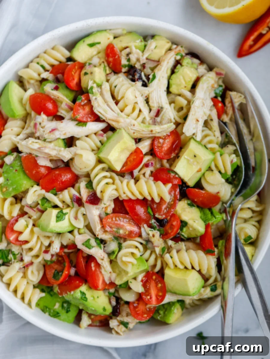 A vibrant bowl of avocado chicken pasta salad, garnished and ready to serve, with a fork and spoon resting inside.