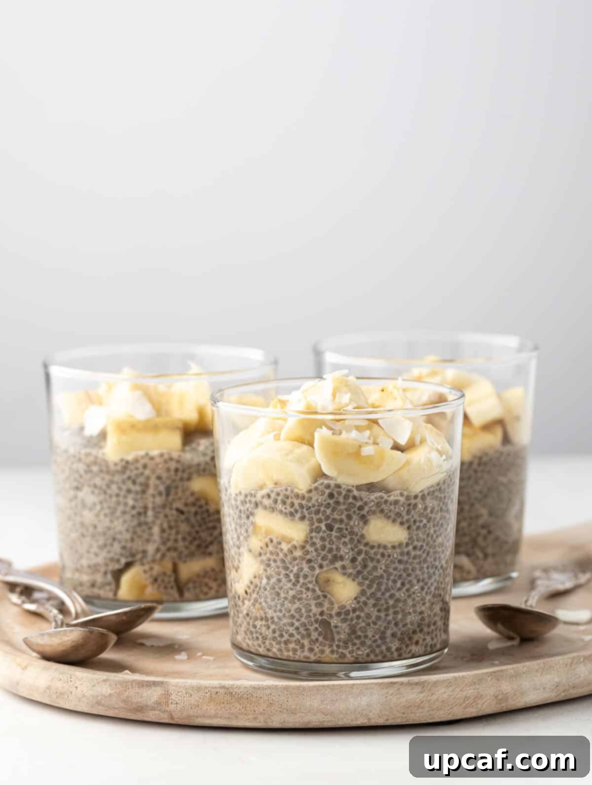 3 cups of banana chia pudding displayed on a wooden board, garnished with fresh banana slices and coconut flakes