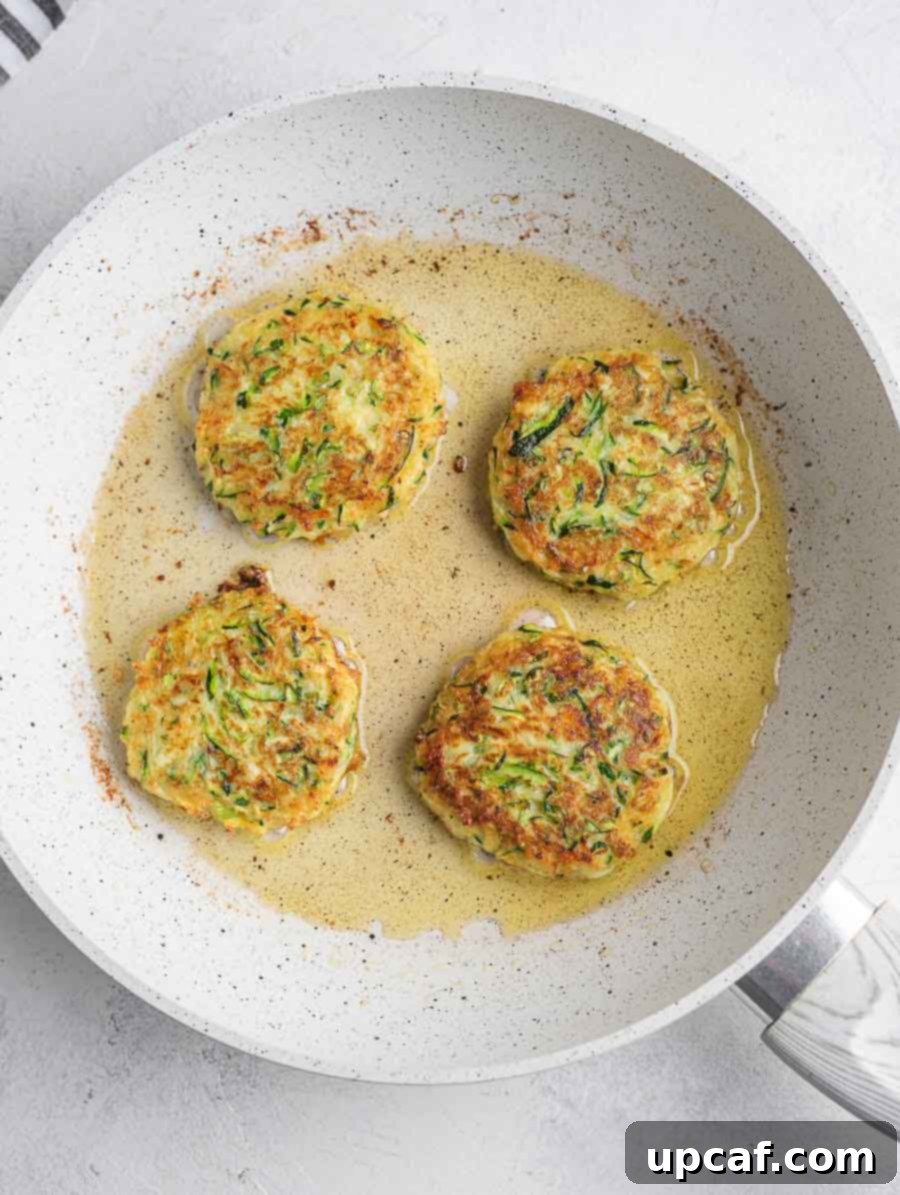Crispy Zucchini Pancakes 7 Several zucchini fritters sizzling and turning golden brown in a hot pan during the frying process.