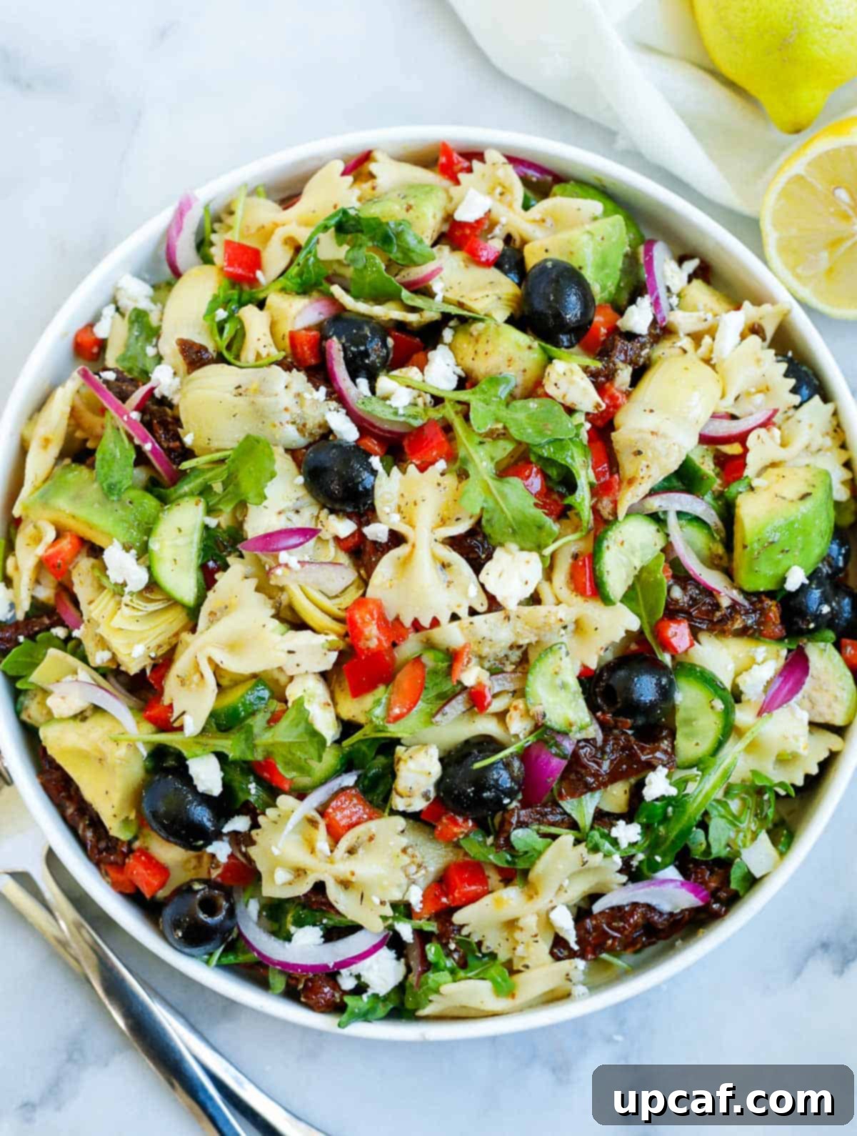 A beautifully presented, fresh lemon herb Mediterranean pasta salad in a white serving bowl, with a side of lemon slices.