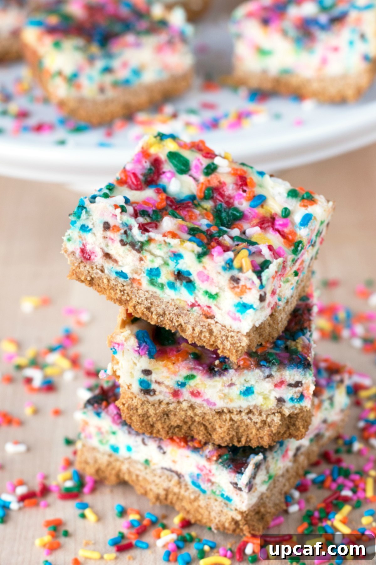 Three funfetti cheesecake bars stacked on a table and surrounded by spirnkles