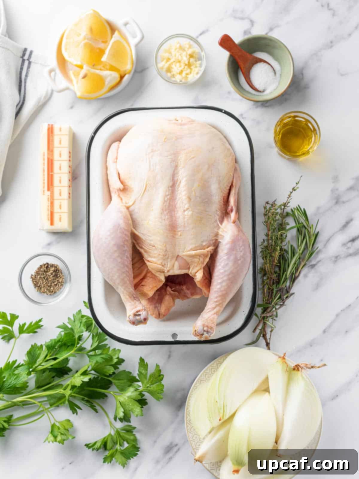 A spread of fresh ingredients including a whole chicken, herbs, garlic, lemon, onion, and butter, ready for roasting.