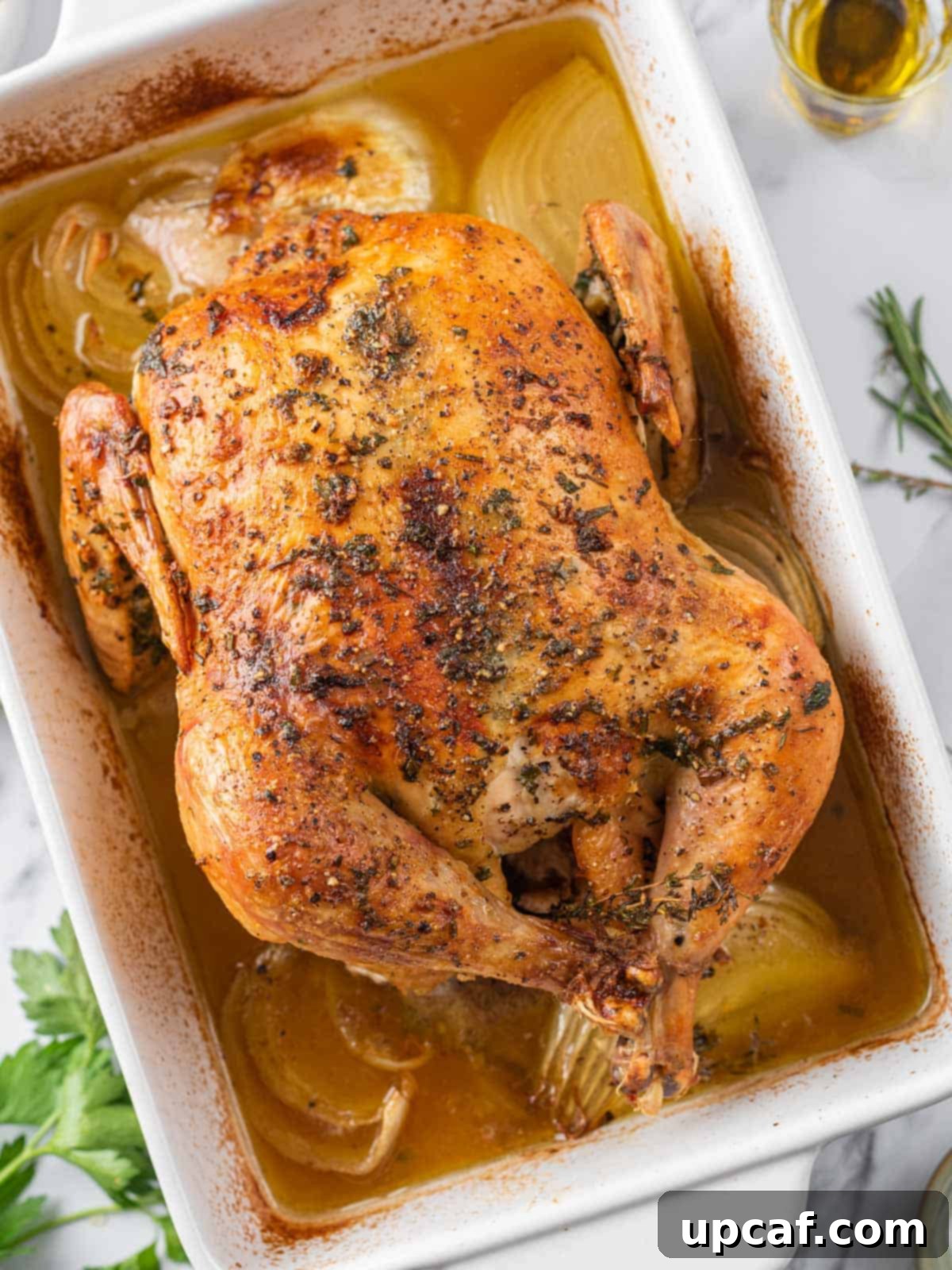 A whole roasted chicken, perfectly golden, baking in a dish with chicken broth.