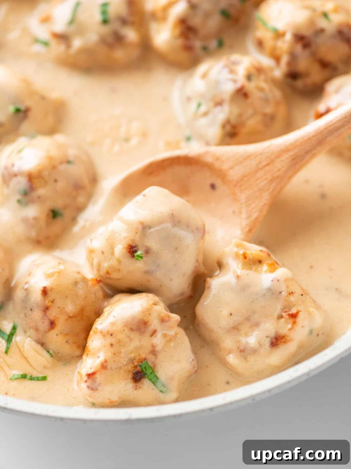 A spoon scooping up a Swedish chicken meatball from a plate, coated in creamy sauce.