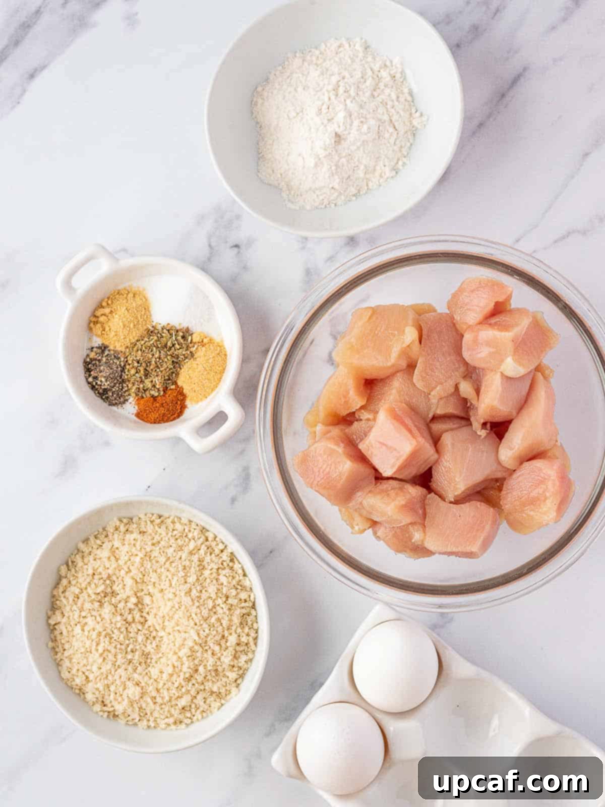 Various ingredients laid out for making popcorn chicken, including chicken, flour, eggs, and spices.