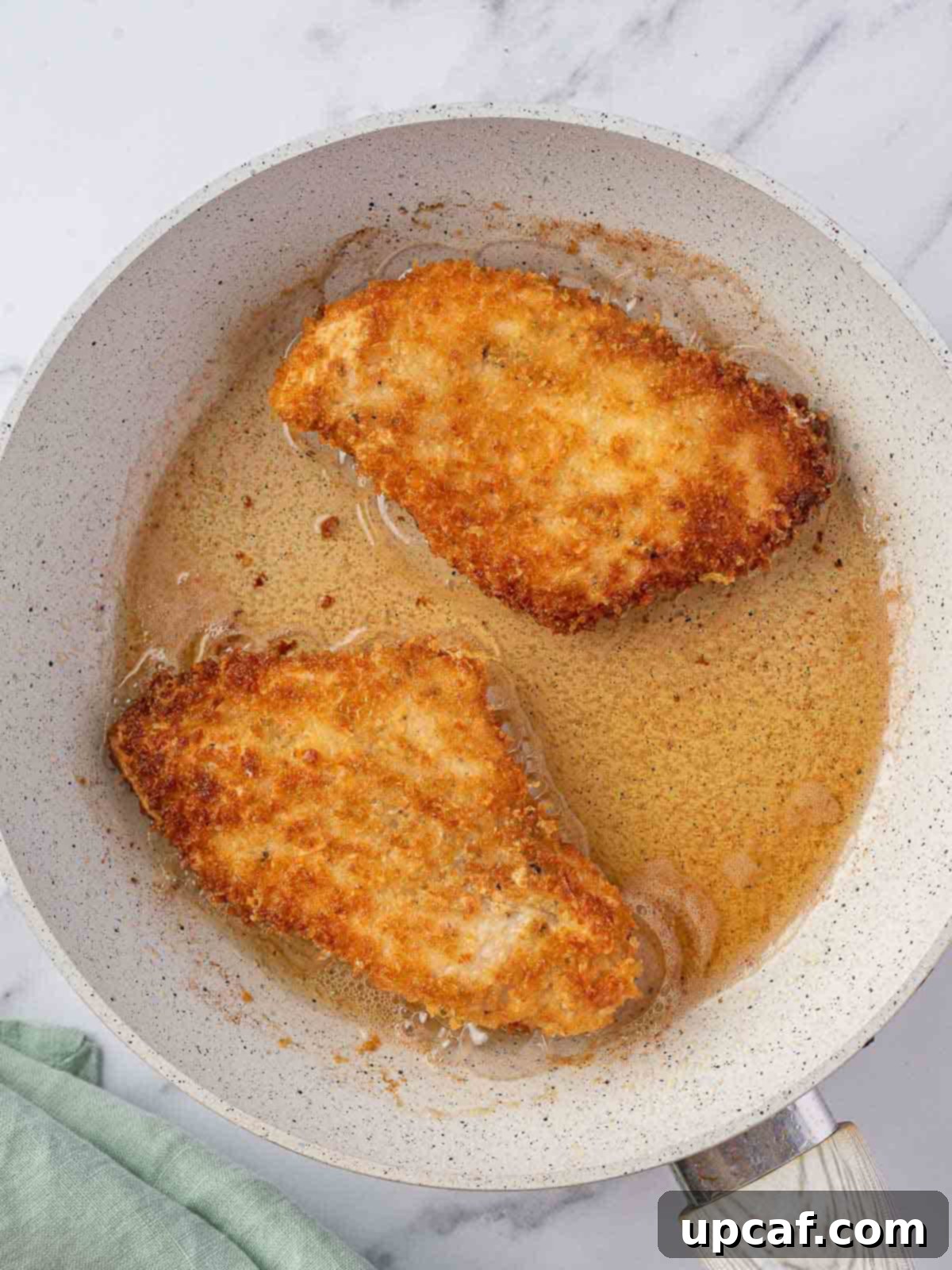 Two pan-fried chicken breasts sizzling in a skillet.