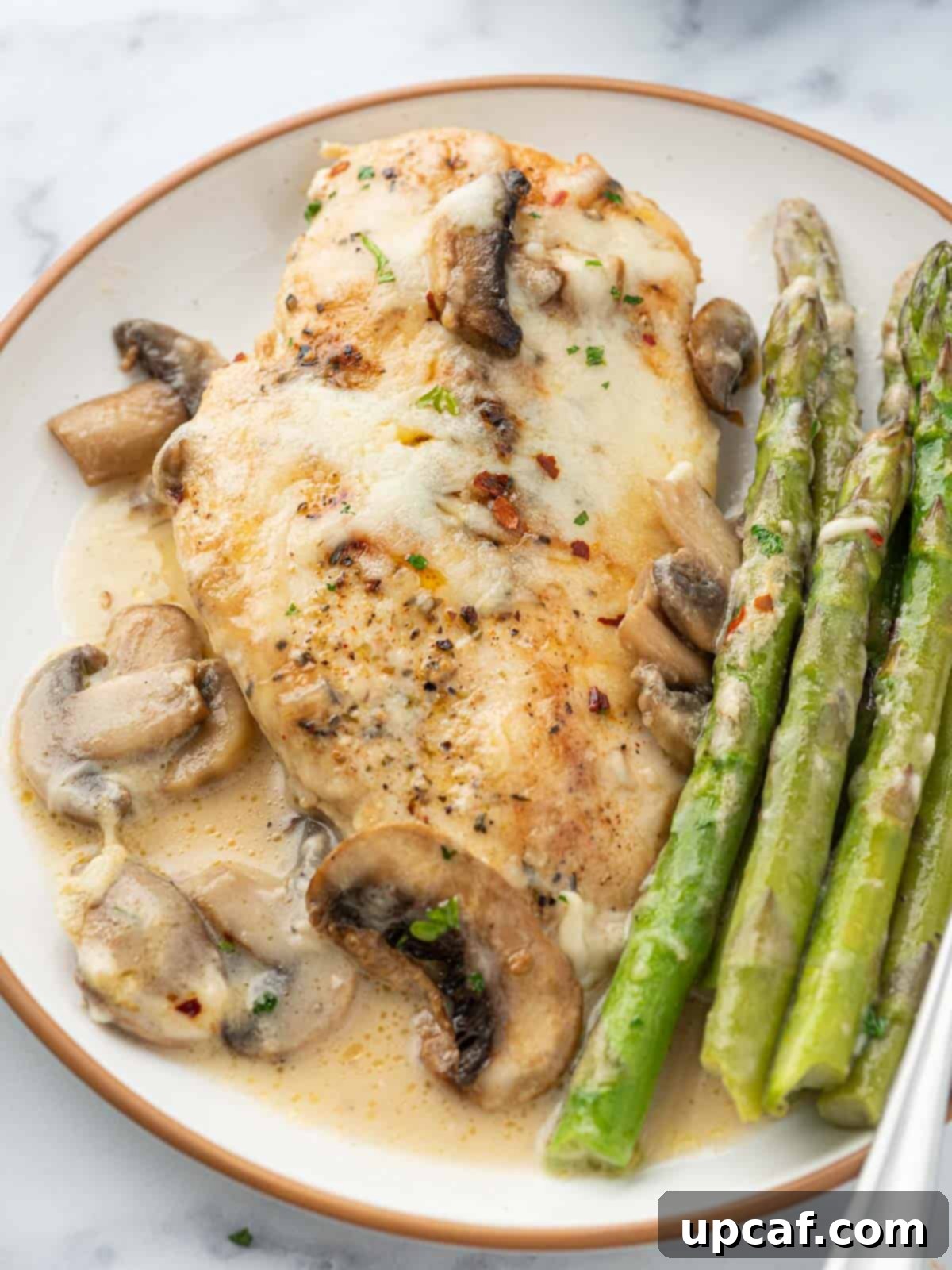 A single serving plate of Cheesecake Factory Chicken Madeira, beautifully plated with tender chicken, mushrooms, and asparagus in a creamy, cheesy sauce, garnished with fresh parsley.