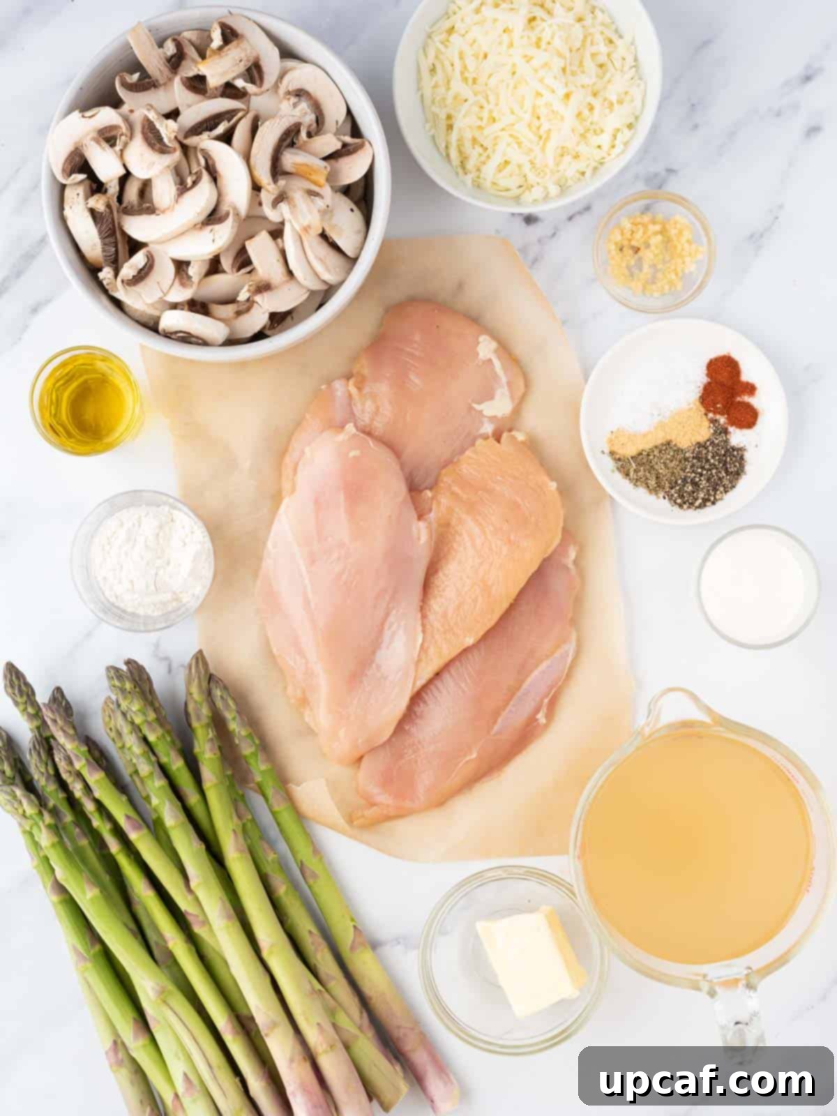Various fresh ingredients laid out on a kitchen counter, including raw chicken breasts, green asparagus spears, whole mushrooms, garlic, and various seasonings, ready for making chicken madeira.