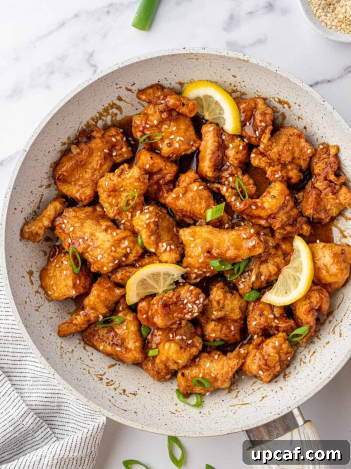 Skillet full of golden lemon chicken topped with vibrant green onions, sesame seeds, and three fresh lemon wedges, ready to be served.