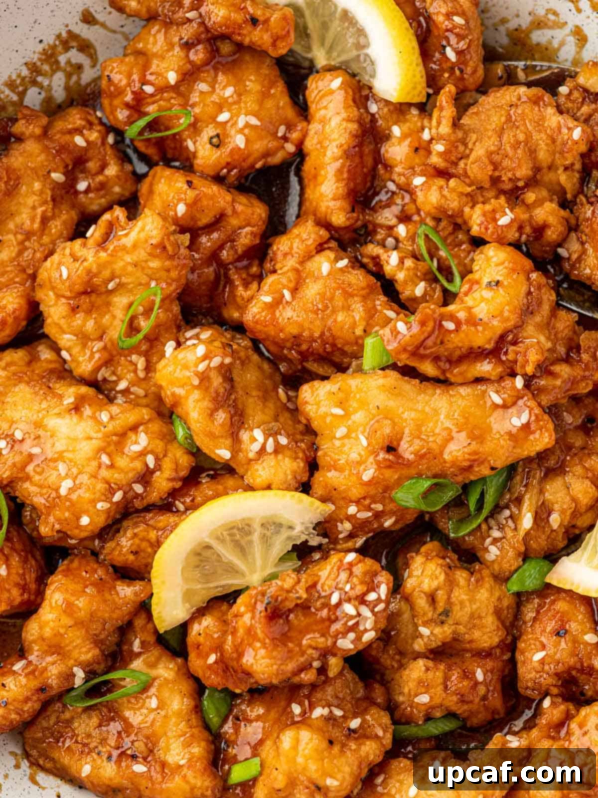 Close-up shot of crispy Chinese lemon chicken topped with shiny sesame seeds and a fresh lemon wedge, highlighting its appetizing texture.