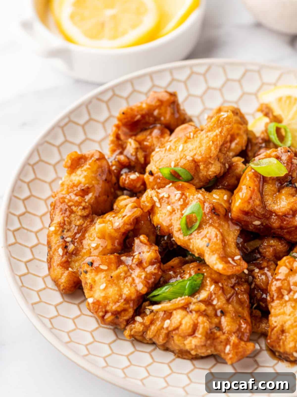 Close-up of golden brown chicken pieces coated in a glossy lemon sauce, garnished with sesame seeds and green onions on a white plate.