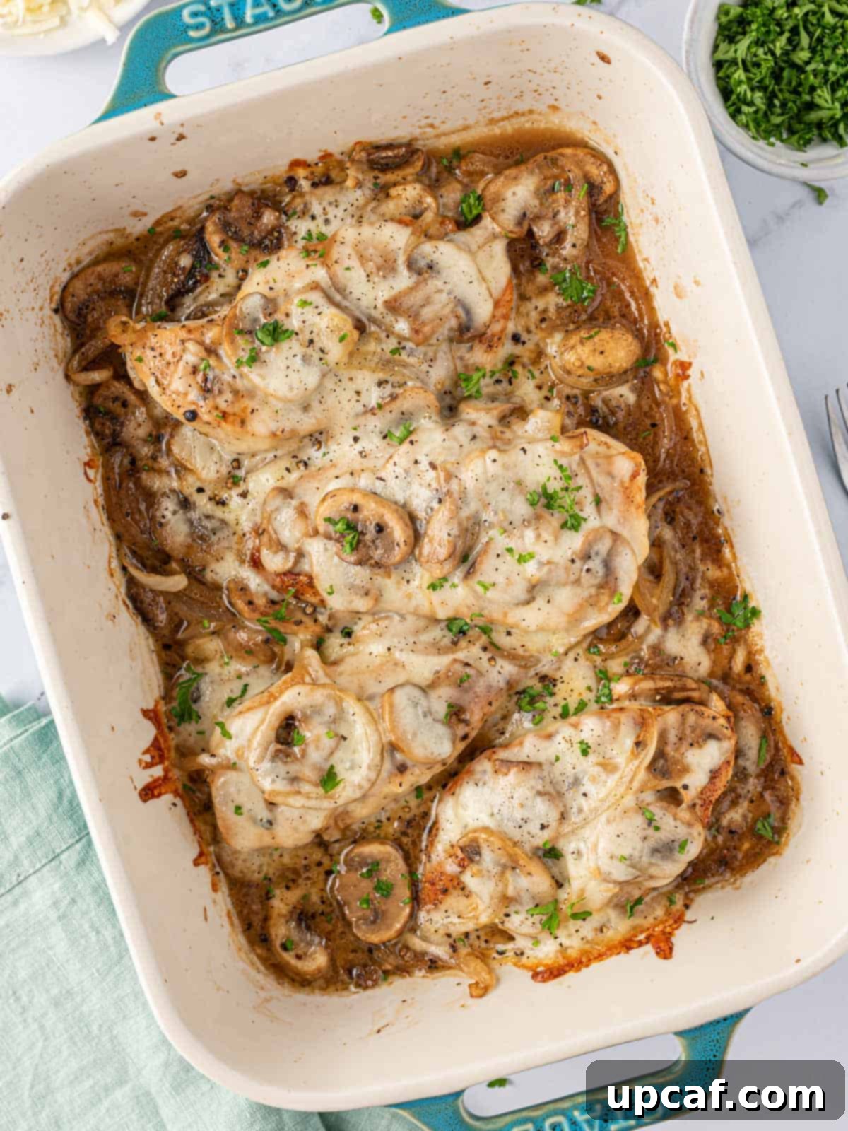 Overhead view of cheesy baked chicken breasts with mushrooms in a blue baking dish, garnished with fresh parsley.