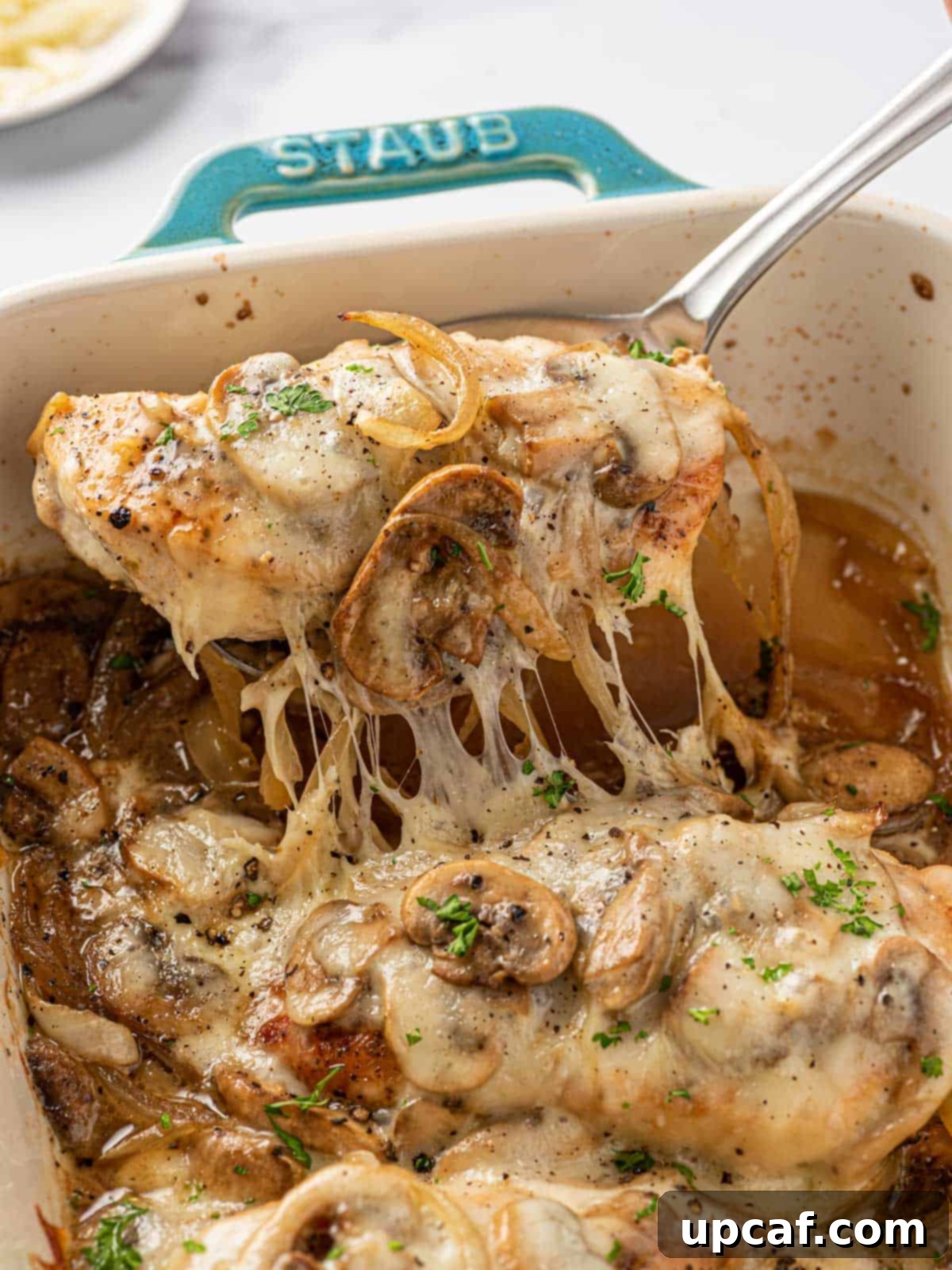 A spoon scooping a piece of cheesy baked chicken breast with mushrooms from the baking dish, showing the gooey cheese and tender chicken.