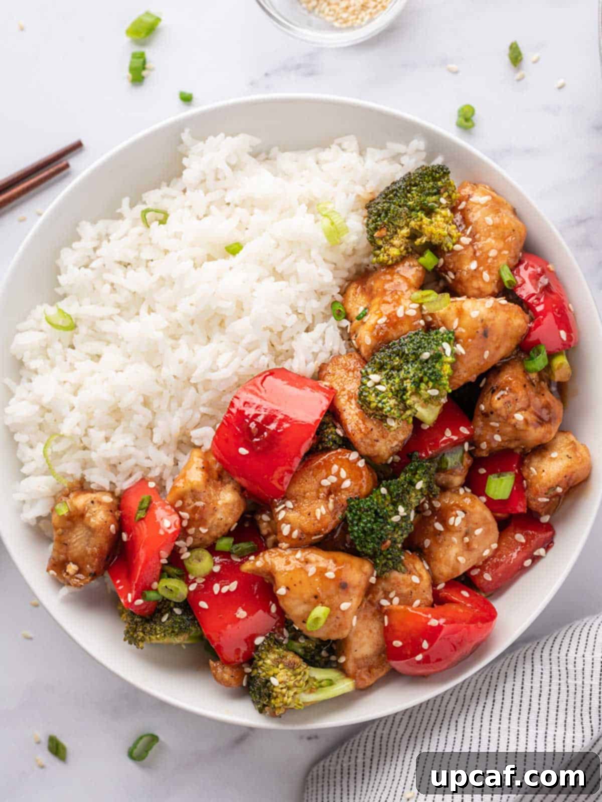 A beautifully presented white bowl filled with rice, topped generously with Teriyaki chicken stir fry, garnished with sesame seeds and chopped green onions.