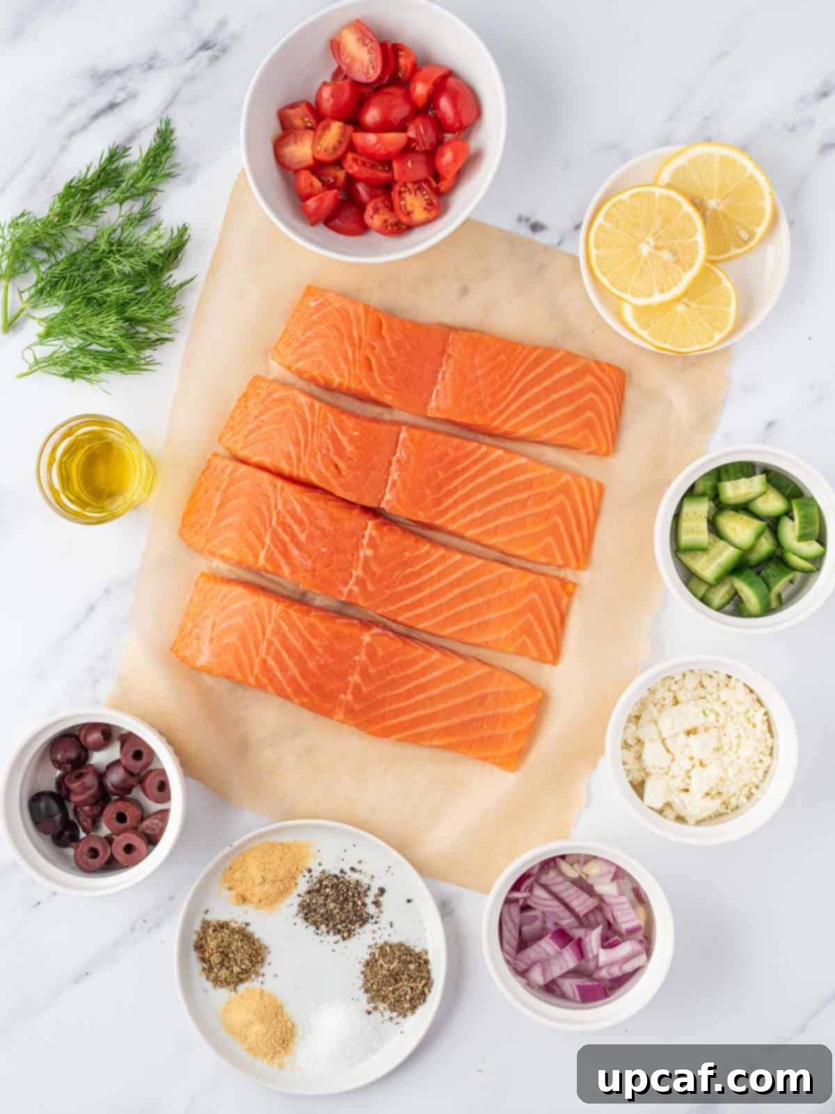 A selection of fresh ingredients including salmon fillets, various herbs, tomatoes, cucumber, and olives, laid out for a Greek salmon recipe.