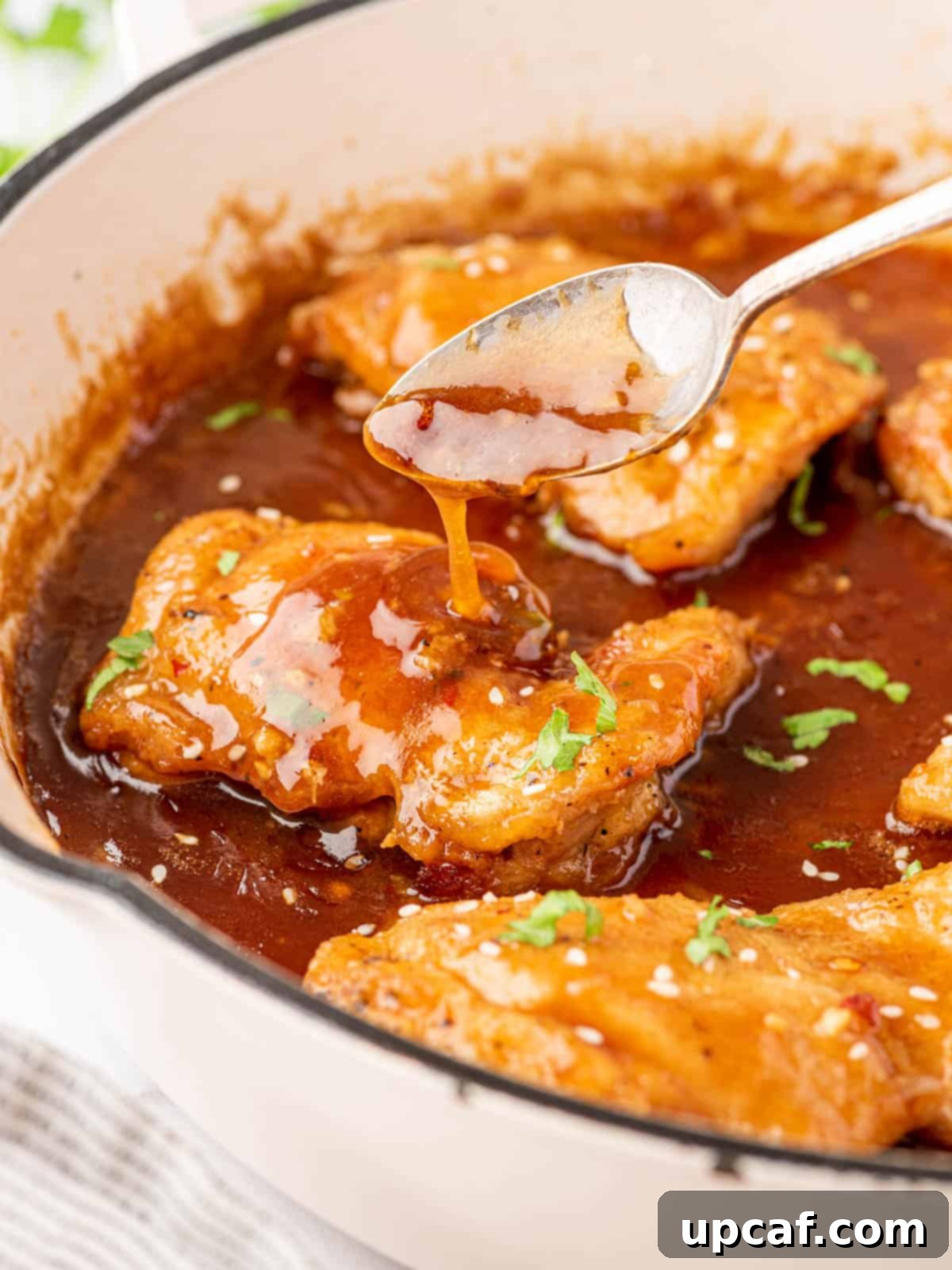 A spoon with a drizzle of honey garlic sauce over a chicken thigh, with a full plate of chicken in the background.