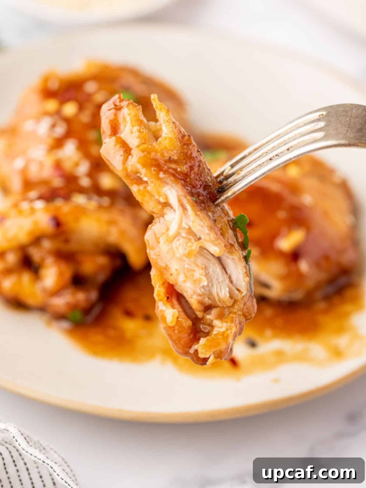 Juicy chicken thigh meat on a fork with a plate of honey garlic chicken in the background, garnished.