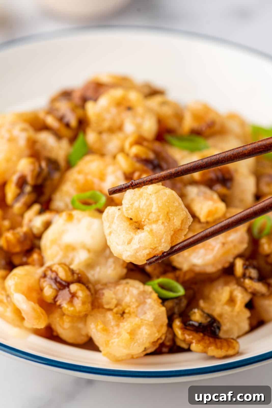 A pair of chopsticks picking up a succulent piece of honey walnut shrimp from a serving bowl, highlighting its crispy texture and creamy coating.