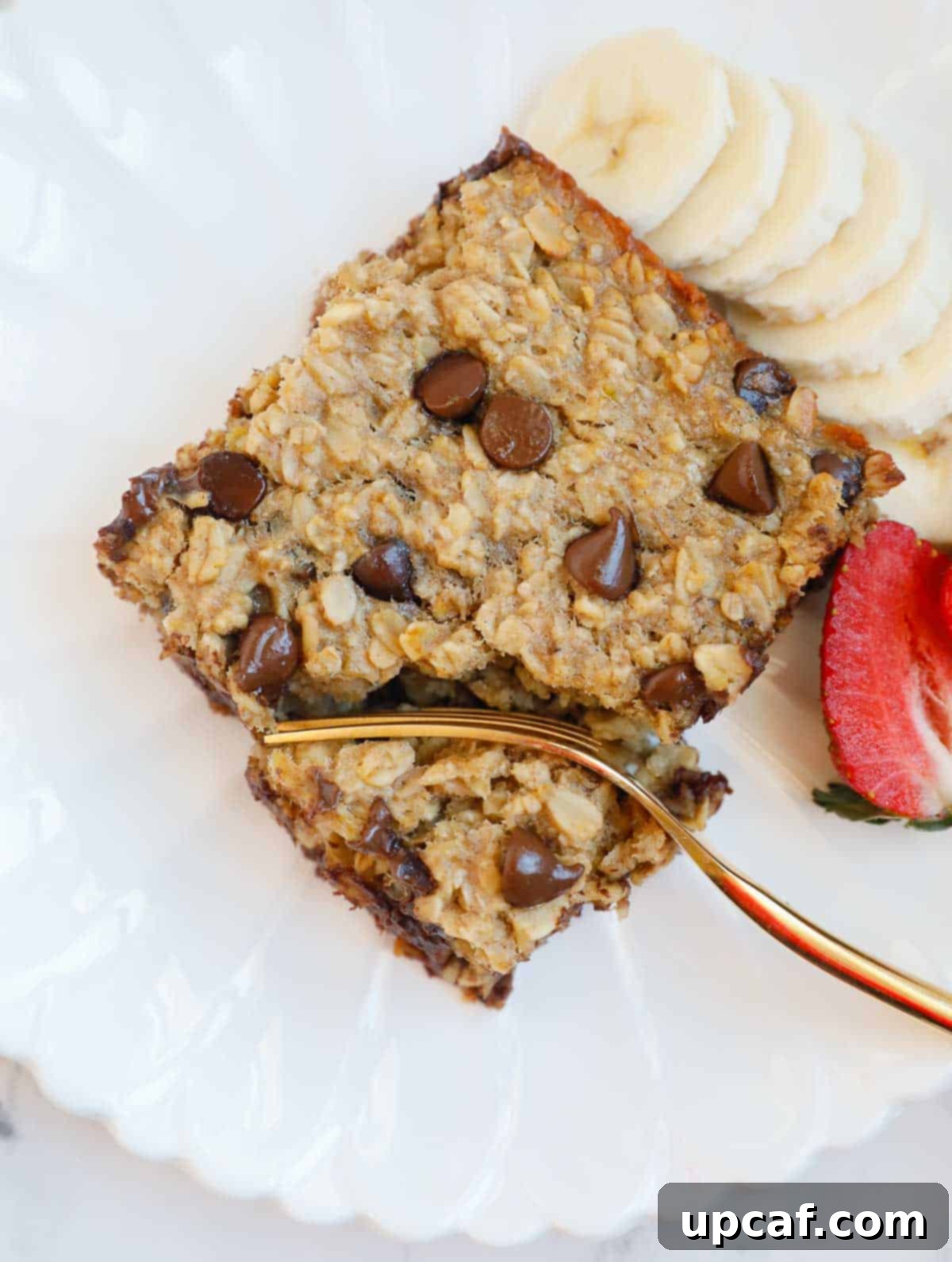 A beautifully arranged plate featuring a square of chocolate chip baked oatmeal, fresh raspberries, sliced bananas, and a fork, illustrating a perfect breakfast.