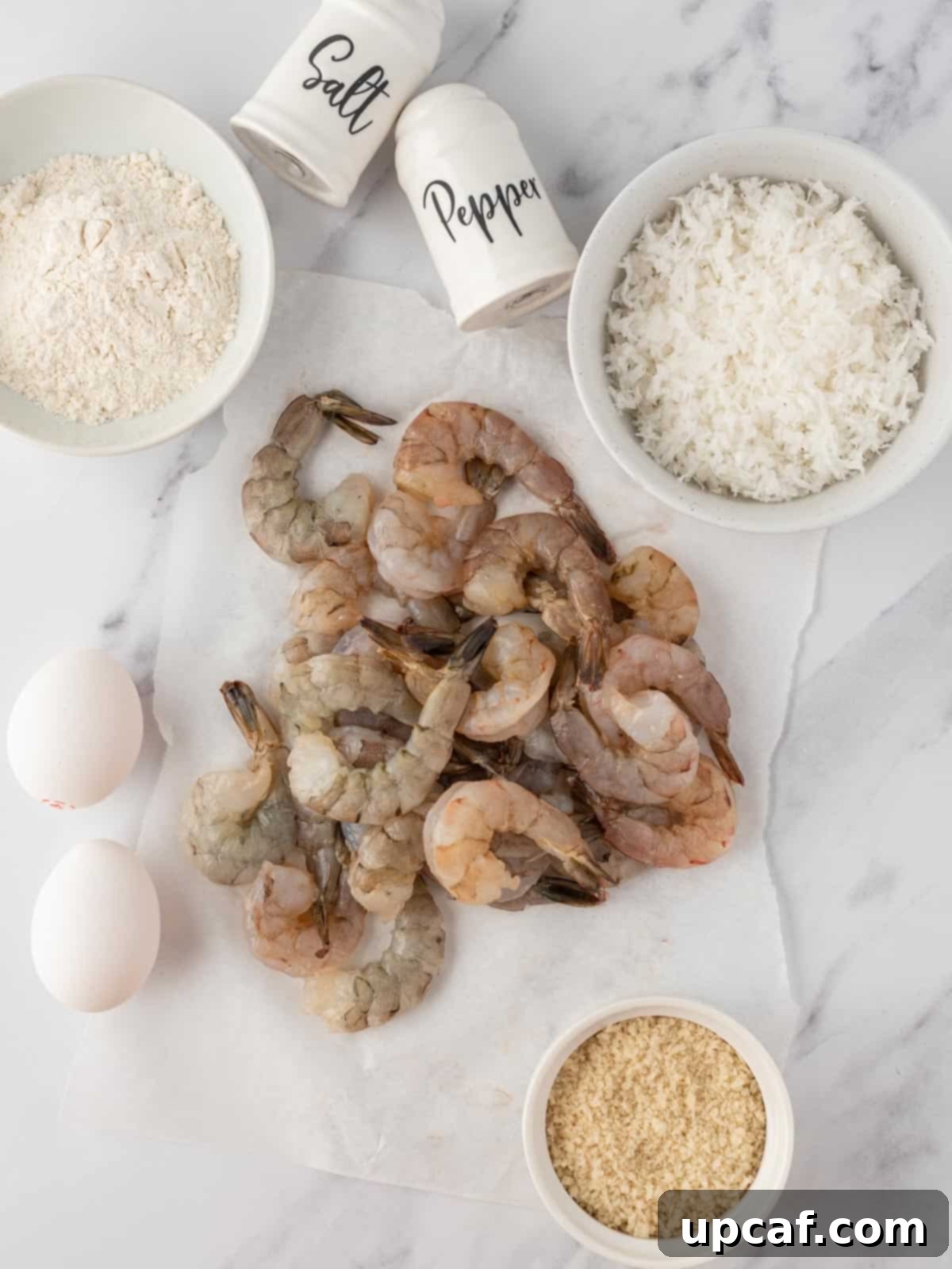 Fresh ingredients laid out for preparing crispy air fryer shrimp, including raw jumbo shrimp, coconut flakes, panko bread crumbs, flour, eggs, and seasonings.