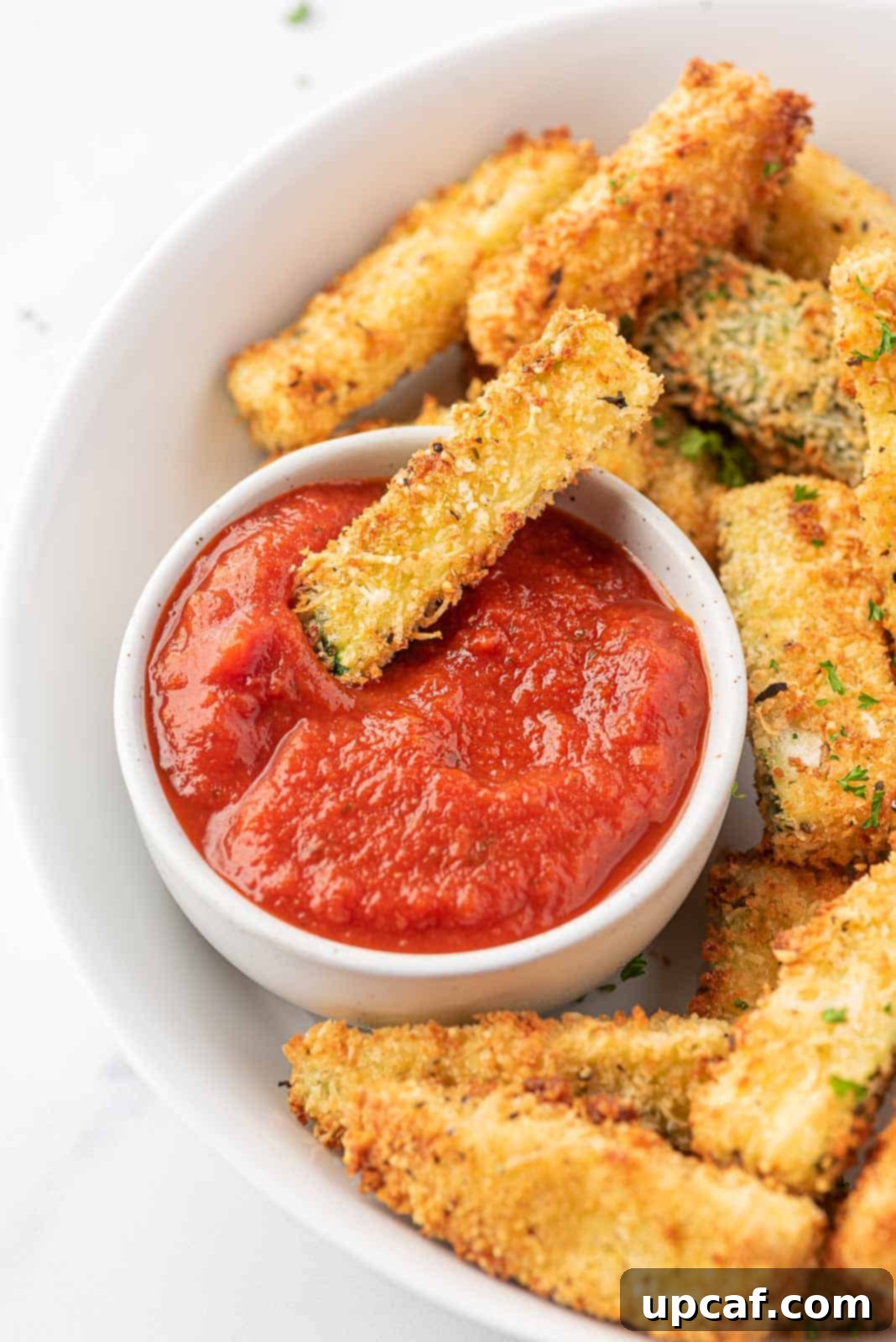 A plate showcasing golden crispy zucchini fries, with one dipped in marinara sauce.