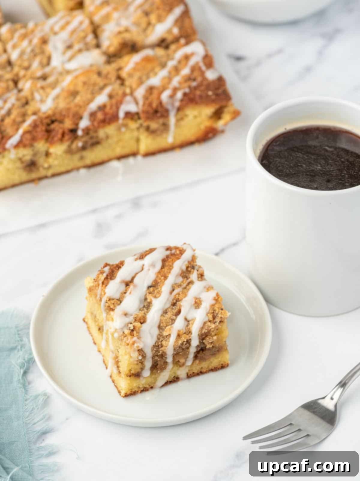 A slice of keto coffee cake on a white plate next to a cup of coffee and a fork. A tray of almond flour coffee cake sits in the background, inviting you to enjoy a guilt-free breakfast.
