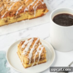 A slice of keto coffee cake on a white plate next to a cup of coffee and a fork. A tray of almond flour coffee cake sits in the background.