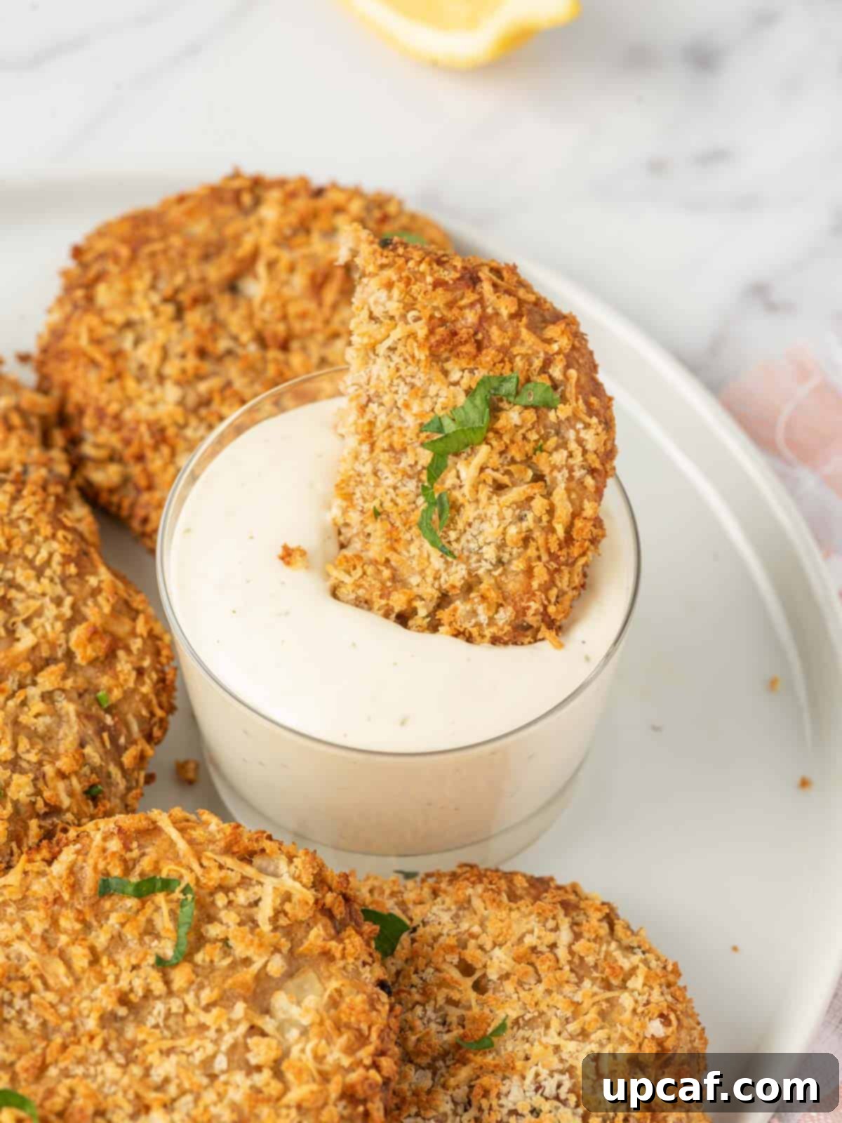 A golden brown air fryer tuna patty being dipped into a creamy white sauce, showing its crispy texture.