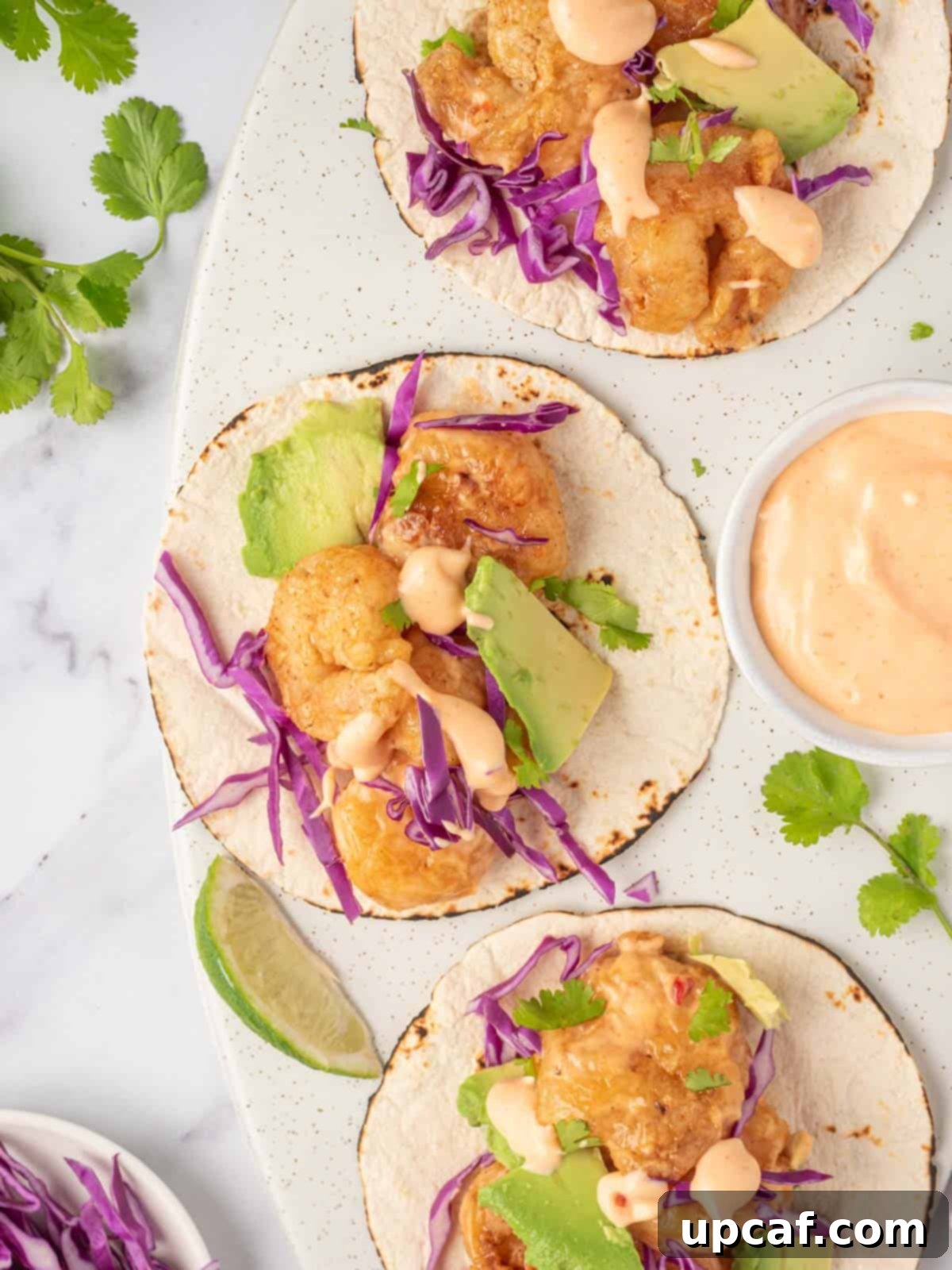 Three bang bang shrimp tacos with dynamite sauce sit on a serving platter, garnished with cilantro and lime wedges.