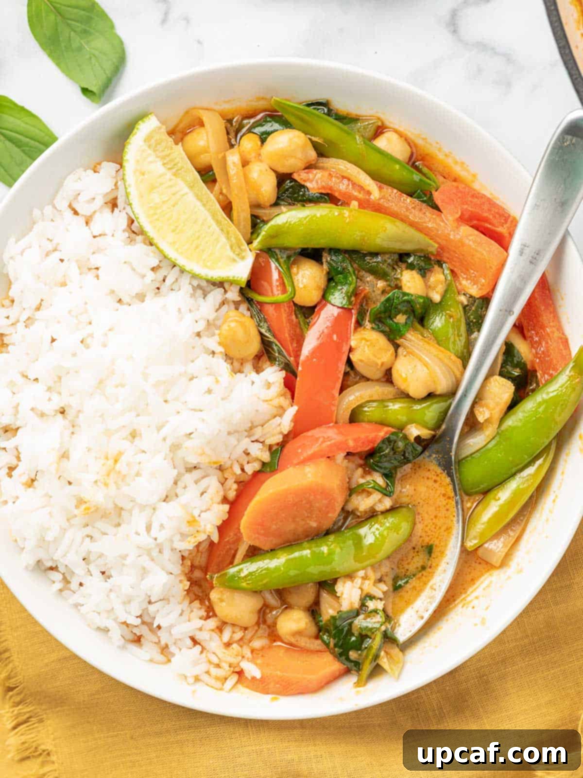 Vibrant Red Thai Plant-Based Curry 9 A generously filled bowl of white rice topped with creamy vegan red curry, vibrant vegetables, and fresh herbs, accompanied by a spoon, highlighting the rich texture and appealing presentation of the meal.